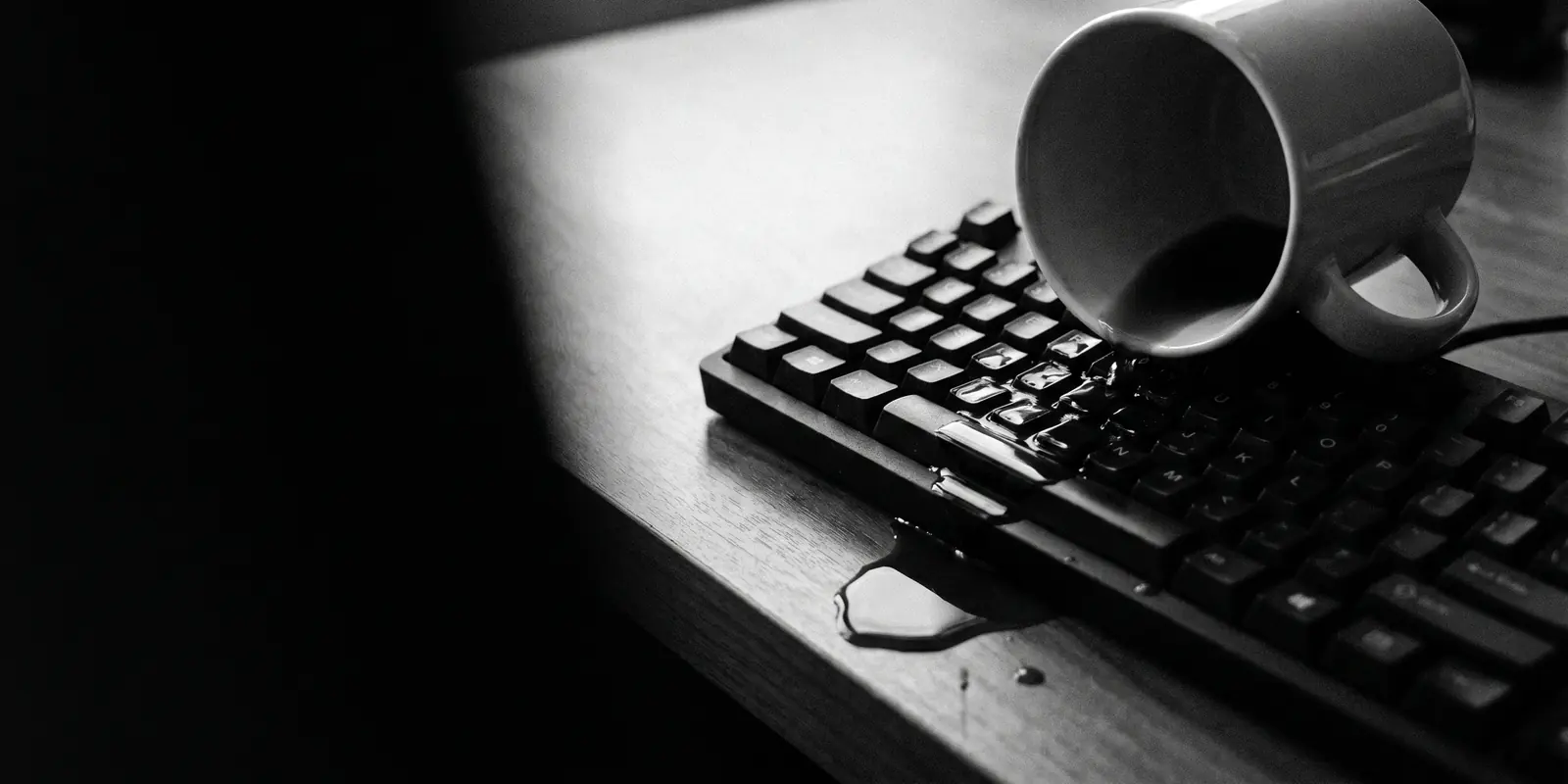 A tipped-over coffee cup lies on a keyboard; coffee spreads across the keys and drips off the desk edge — an image for the everyday mishap.