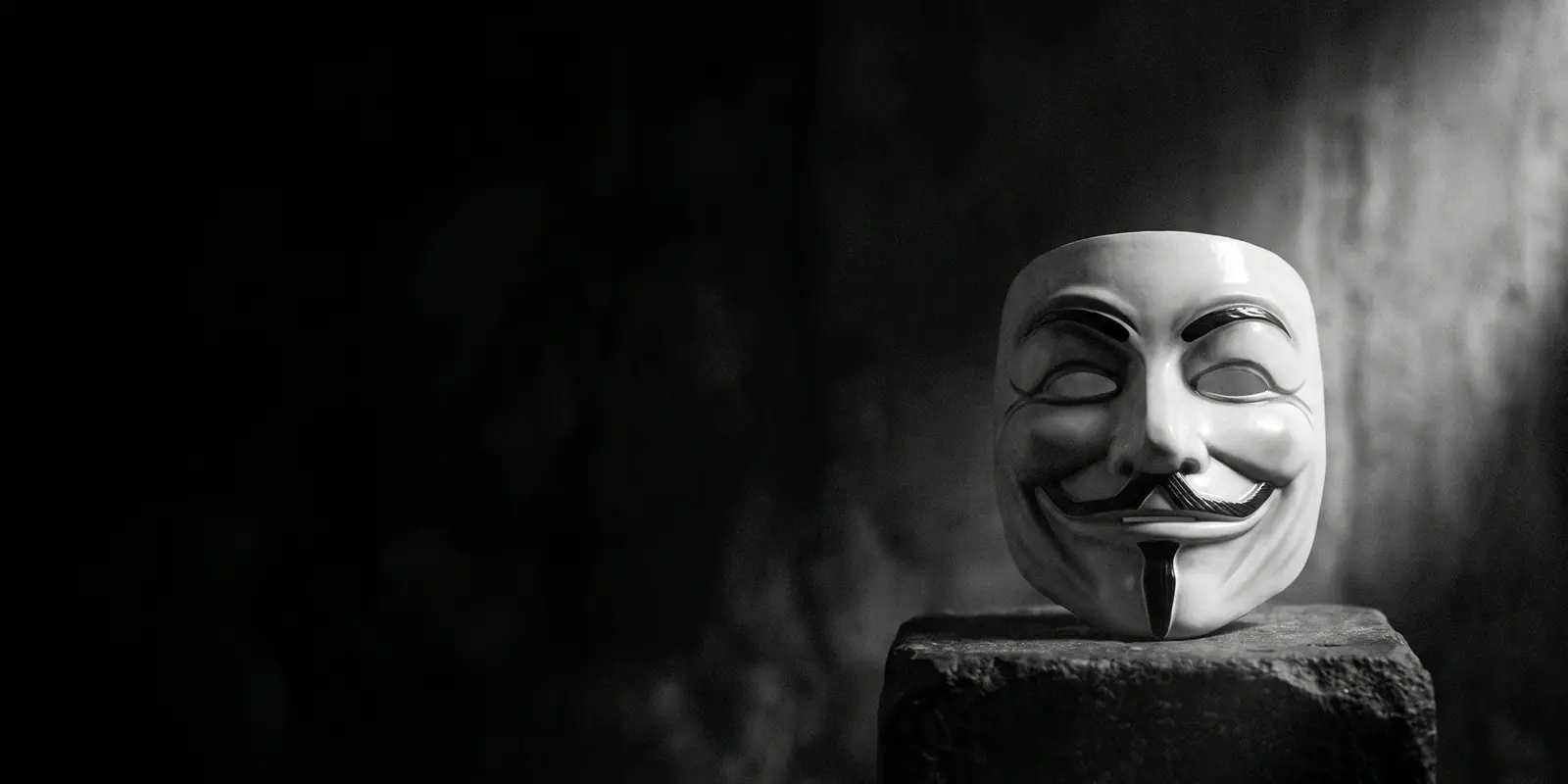 Een Guy Fawkes-/Anonymous-masker staat frontaal op een stenen sokkel in strijklicht tegen een donkere achtergrond — beeld voor de anonieme aanvaller achter een gewone voorkant.